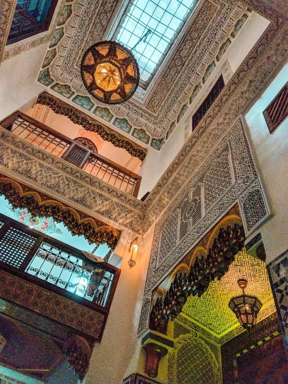 Stunning low-angle view of a traditional Moroccan Riad courtyard in Fes Medina, showcasing intricate hand-carved plasterwork, majestic brass lanterns, and high arched balconies under a natural skylight.