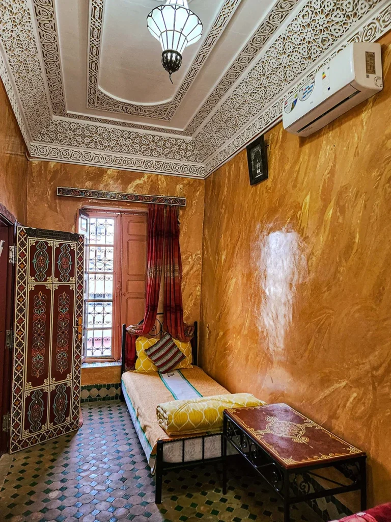 Side view of a cozy bed in a Fez Riad room showing traditional orange Tadelakt walls and hand-painted wooden door decorations.