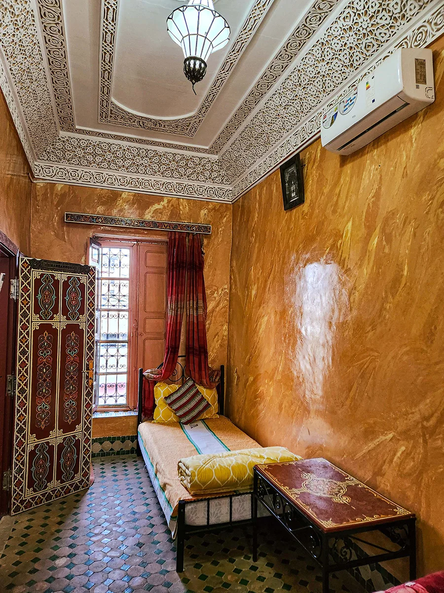 Side view of a cozy bed in a Fez Riad room showing traditional orange Tadelakt walls and hand-painted wooden door decorations.