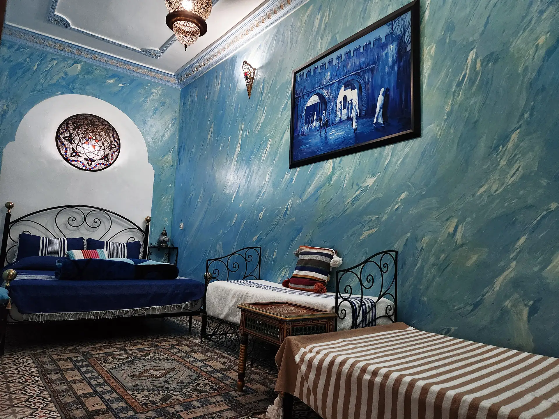 Close-up of a double bed in the Deluxe Suite featuring a traditional stained glass window, blue Tadelakt walls, and a painting of a Moroccan gate.