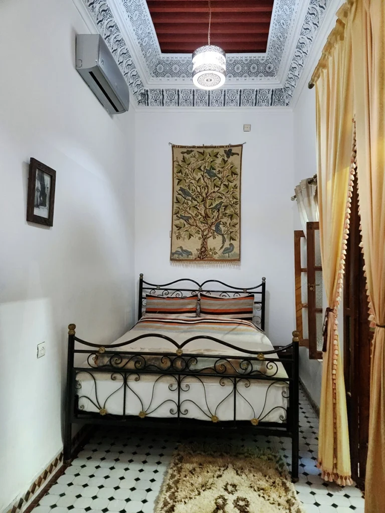 Main bedroom area of the Grand Family Suite in a Fez Riad featuring a double bed, high ornate plaster ceiling, and a traditional tree-themed wall hanging.