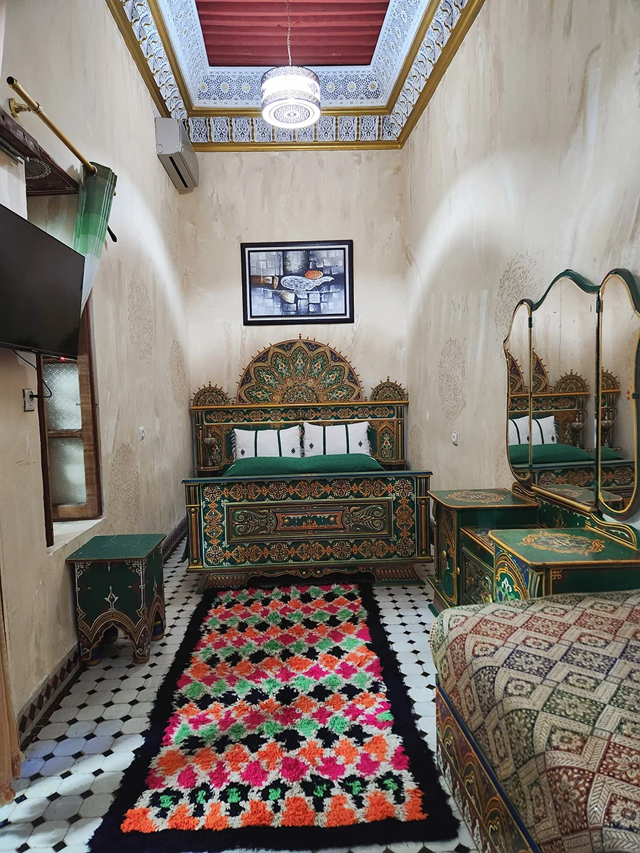 Comprehensive view of the Royal Heritage Suite in Riad Fez featuring a magnificent green hand-carved bed, traditional colorful rug, and authentic Moroccan wall decor.
