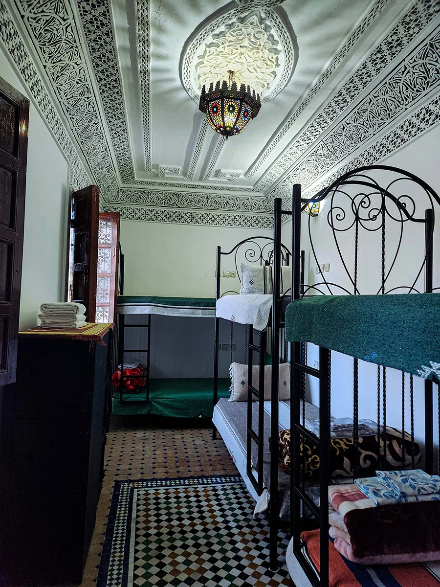 Wide view of a spacious shared group room in a Fez Riad featuring three metal bunk beds for six guests, a beautifully carved white plaster ceiling, and traditional tiled flooring.