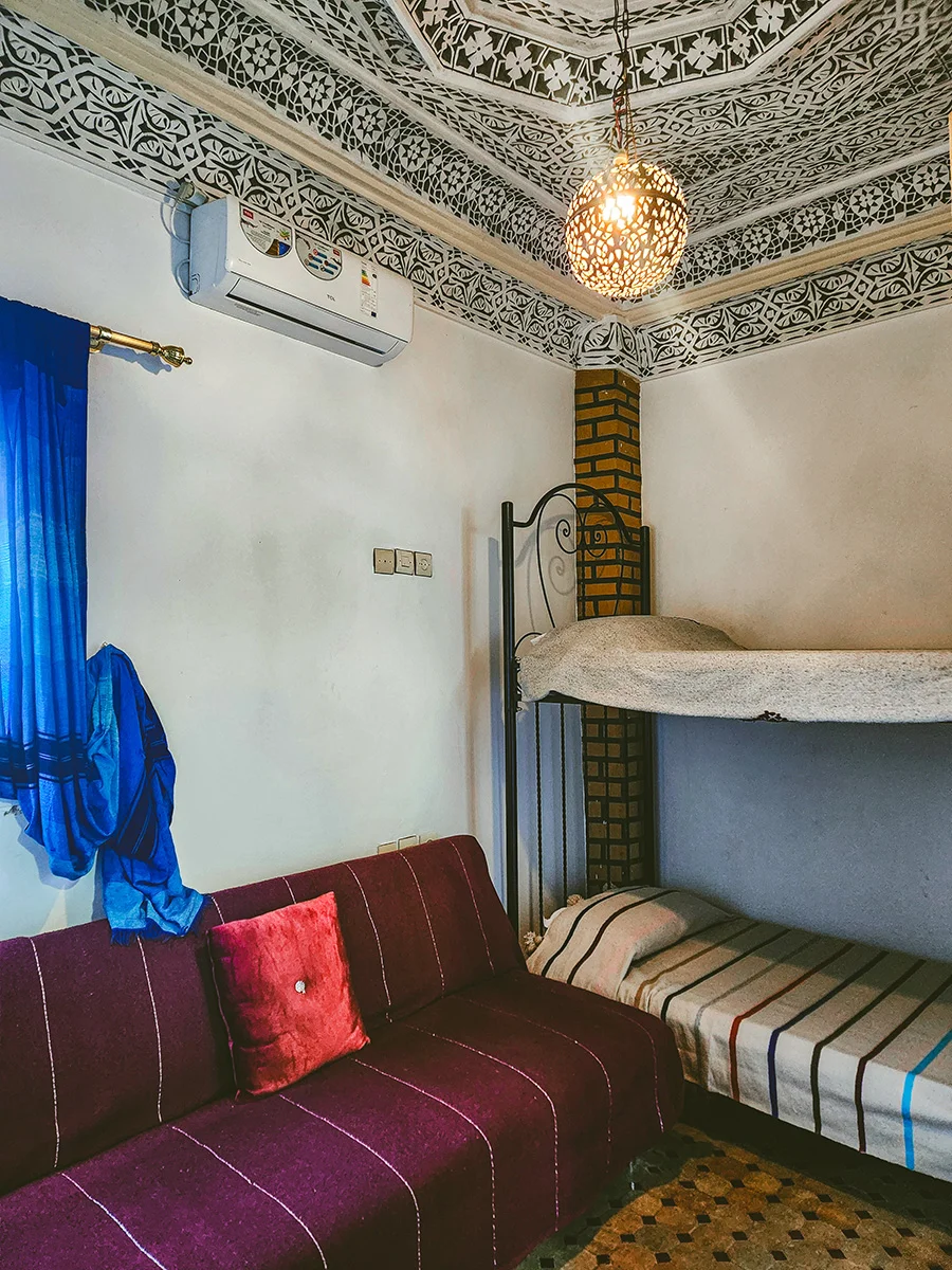 Standard Economy Room in a Fez Riad featuring a sturdy bunk bed, a purple sofa bed, and detailed traditional Moroccan plasterwork on the ceiling.