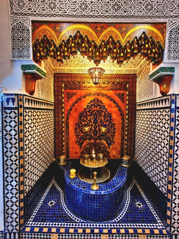 An authentic Moroccan wall fountain (Sekkaya) in a Fez Riad, decorated with intricate blue and white Zellije tiles, hand-carved plasterwork, and a traditional brass tea service. Authentic Riad Experience Fez - Riad Blida Courtyard